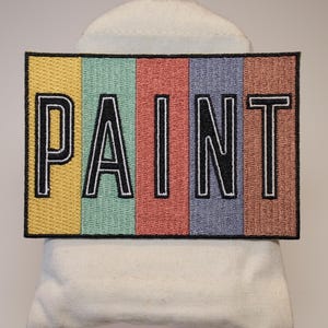 Puede incluir: Un parche rectangular con la palabra "PAINT" bordada en negro, contorneada en blanco, sobre un fondo colorido. El fondo está dividido en franjas verticales amarillas, verde claro, rojas, azules y marrones. El parche se muestra sobre una superficie blanca.