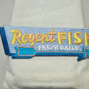 Könnte beinhalten: Gestickter Aufnäher mit den Worten "Regent FISH FRESH DAILY" in gelben und orangefarbenen Buchstaben. Der Aufnäher hat einen hellblauen Hintergrund mit einem blauen Pfeil, der nach rechts zeigt. Der Aufnäher ist rechteckig und hat einen blauen Rand.