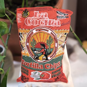 Puede incluir: Un parche bordado rojo y verde de una bolsa de chips de tortilla con el texto "La Cocina Tortilla Chips" y una imagen de dibujos animados de una persona haciendo tortillas.