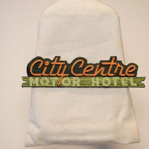 以下が含まれることがあります： オレンジと緑色の「City Centre MOTOR HOTEL」という文字が刺繍されたパッチ。パッチは白い布に取り付けられています。パッチはレトロなデザインです。