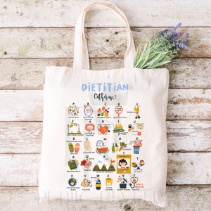 May include: A white tote bag with a blue and white design that says "Dietitian Catherine". The bag has a colorful alphabet chart with illustrations and words related to nutrition and health.