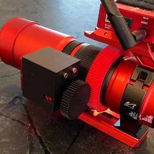 May include: A red and black camera lens with a gear system and a black box attached. The lens has a red housing and a black focus ring. A red component with a Wi-Fi and reset button is also visible.