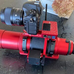 May include: A black digital camera with a large lens is mounted on a red telescope and support system. The setup includes a red control box with ports and a black antenna. The equipment is designed for astrophotography.