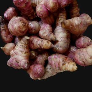 May include: A close-up of a pile of Jerusalem artichokes. The tubers have a knobbly, irregular shape and a mix of colours, including shades of pink, brown, and beige. The background is a solid black.