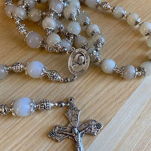 May include: A rosary with light gray and white beads and ornate silver-colored metal accents. The rosary includes a crucifix and a medal with an image of Saint John Paul II.