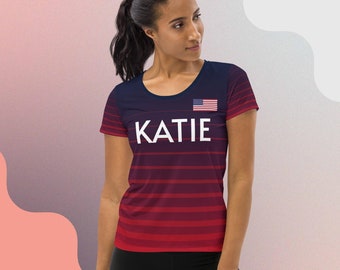 May include: A navy blue and red athletic t-shirt with the name "KATIE" in white block letters. The shirt features horizontal stripes and a small American flag on the upper right chest.