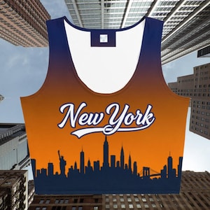 May include: A sleeveless crop top with an orange and blue gradient design. The front displays the text "New York" in white script above a dark blue silhouette of the New York City skyline, including the Statue of Liberty and the Brooklyn Bridge.