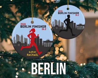 May include: Two round Christmas ornaments with a running theme. One shows a red runner with the text "2026 Berlin Finisher 26.2" and "4:49:41 RACHAEL". The other shows a black runner with the same text. The word "BERLIN" is at the bottom.