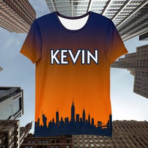 May include: A short-sleeved t-shirt with a navy blue to orange gradient design. The name "KEVIN" is printed in white block letters. A dark blue city skyline is featured at the bottom of the shirt, with iconic buildings.