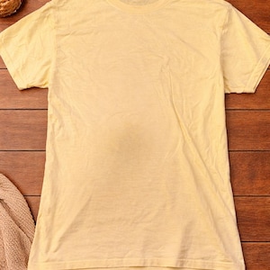 May include: A pale yellow crew neck t-shirt with short sleeves. The shirt is made of a soft material and is slightly wrinkled. The shirt is displayed on a wooden surface with a straw hat and other items in the background.