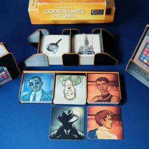 May include: A board game set, "Codenames Pictures", with various components. The game includes cards with illustrations, a game board, and a box. The cards feature images of a frying pan, a building, and people. The box is yellow and black.