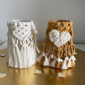 Peut inclure: Deux bougeoirs en macramé, l'un blanc et l'autre jaune moutarde, avec des pompons blancs et des glands. Les bougeoirs sont sur une surface dorée avec des cœurs en bois blanc.
