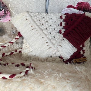 Pode incluir: Uma bolsa de crochê branca e bordô com detalhes em franjas e uma alça trançada. A bolsa tem uma etiqueta de madeira com o texto "Feather Art".