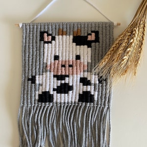 Peut inclure: Une tapisserie murale tissée en gris et blanc avec un motif de vache. La vache est réalisée avec du fil blanc, noir, brun et rose. La tapisserie a des franges grises en bas.