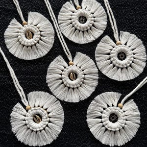 Puede incluir: Seis tapices de macramé blancos con un diseño circular y una pequeña concha de mar en el centro. Cada tapiz tiene una cuenta de madera y un cordón blanco.