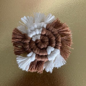 Puede incluir: Un posavasos de macramé hecho a mano con un diseño en espiral. El posavasos presenta secciones alternas de cordón de algodón anudado marrón y blanco, con una franja de borlas alrededor del borde exterior. El posavasos es circular y descansa sobre una superficie dorada.