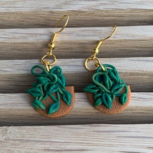 May include: A pair of gold dangle earrings with green and brown clay charms shaped like potted plants. The plants have green leaves and vines.