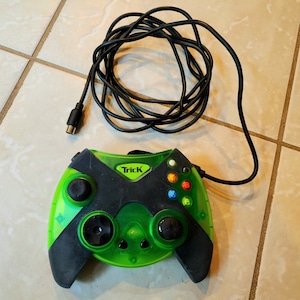 Puede incluir: Un mando de videojuego verde y negro con la palabra "Trick" impresa en él. El mando tiene seis botones y dos joysticks.
