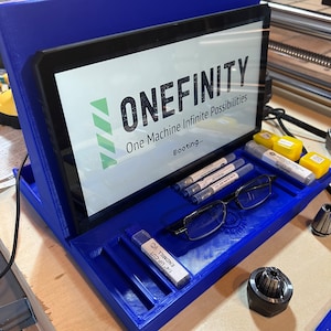 Puede incluir: Un soporte de impresión 3D azul con una pantalla que muestra el texto "ONEFINITY One Machine Infinite Possibilities Booting..." El soporte tiene una bandeja con un par de gafas, una pequeña botella etiquetada "1/4" Endmill", y otros objetos pequeños.