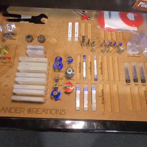 May include: A wooden organiser with various tools and supplies for woodworking. The organiser has slots for holding different sizes of router bits, drill bits, and other tools. The organiser also has a label that says "SANDER CREATIONS".