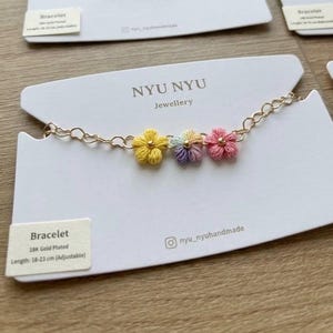 May include: An 18K gold plated chain bracelet featuring three small, embroidered flower charms in yellow, a multi-colored pastel blend, and pink. The bracelet is displayed on a white card labeled NYU NYU Jewellery with a length of 18-23 cm.