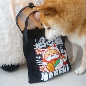 May include: Black tote bag with a white cartoon cat wearing a red and gold outfit. The cat is surrounded by pink flowers and the text "Maneki" in white. The bag has a white Japanese character on the top left corner and the text "Kyu Kyu" on the bottom right corner.