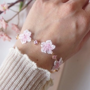 Peut inclure: Un bracelet délicat avec des fleurs crochetées roses et de petits cristaux roses sur une chaîne dorée.