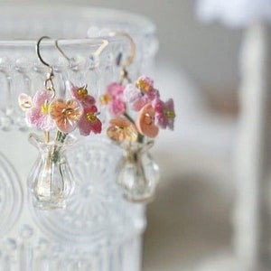 May include: A pair of delicate earrings featuring miniature bouquets of pink and orange flowers in tiny glass vases. The earrings have gold-colored hooks and are set against a clear, textured glass background.