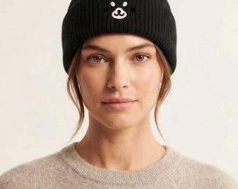 May include: A black ribbed knit beanie featuring a small, white embroidered bear face graphic on the front cuff. The hat has a folded brim design and is constructed from a solid black yarn, showcasing a simple, clean aesthetic for headwear.