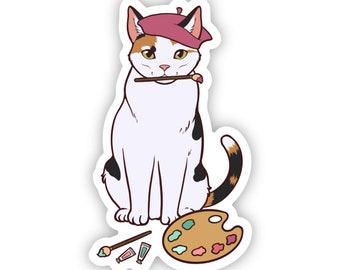 May include: A calico cat wearing a pink beret holds a paintbrush in its mouth and sits next to a paint palette with paint tubes and brushes.
