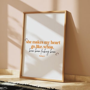 May include: A framed print with a white background and a quote in orange text that reads "she makes my heart go, like, whoa. boom boom fucking boom". The quote is followed by "Binding 13" in black text.