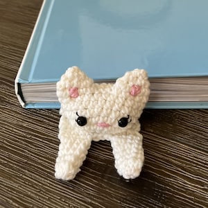 Könnte beinhalten: Handgefertigter, weißer Häkel-Lesezeichen-Hase mit rosa Akzenten und schwarzen Augen. Der Hase liegt auf einem hellblauen Buch, mit einem grauen Blumentopf im Hintergrund. Das Lesezeichen ist ein einzigartiger, handgefertigter Artikel.