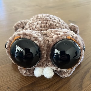 Puede incluir: Un juguete de peluche de color marrón tejido a crochet con grandes ojos negros y una boca blanca. El juguete tiene una textura peluda y está hecho con un hilo suave.