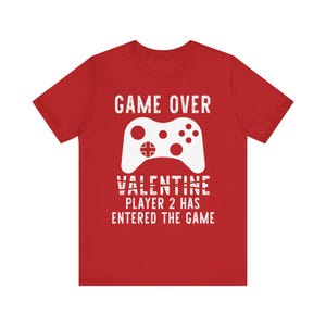 May include: Red t-shirt with a white graphic of a video game controller and the text "GAME OVER VALENTINE PLAYER 2 HAS ENTERED THE GAME". The shirt is made of a soft material.