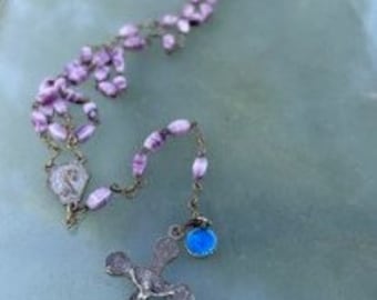 May include: A rosary with light purple beads and a silver crucifix. The crucifix features a detailed depiction of Jesus Christ. The rosary also includes a small, blue charm and a decorative centerpiece.