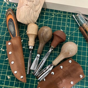 Puede incluir: Un conjunto de cinco herramientas de talla de madera con fundas de cuero. Las herramientas tienen mangos de madera y cuchillas de metal. Las fundas son de cuero marrón con un diseño decorativo.