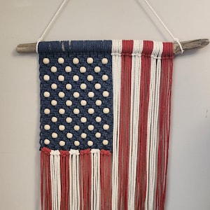 Puede incluir: Un tapiz de pared con la bandera estadounidense hecho a mano con hilo azul, rojo y blanco. La bandera presenta un patrón de estrellas blancas sobre un fondo azul y rayas rojas y blancas. La bandera está unida a un trozo de madera a la deriva.