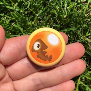 May include: A yellow button with a brown cartoon fish on an orange background. The fish has a large eye and a small mouth.