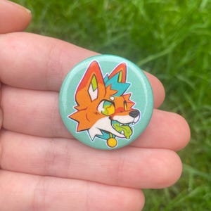 May include: A circular badge featuring a cartoon fox design. The fox is depicted with orange and white fur, green and yellow eyes, and a green tongue. The badge has a light teal background and a gold collar.