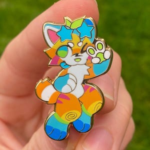 May include: A colourful enamel pin depicting a cartoon cat character with a gold outline. The cat has a white face, orange and yellow body, and blue and green accents. The pin is held in a hand against a blurred green background.