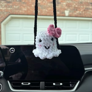 May include: A handmade crochet ghost in white, with a pink bow, black eyes, and a smiling face. It hangs from a black string, intended as a car charm. The background shows a car dashboard and a brick wall.