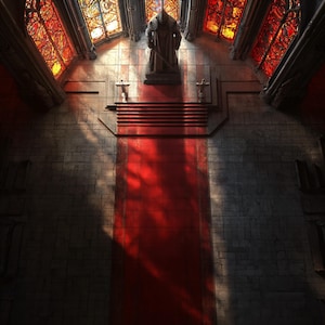 Può includere: Interno di una cattedrale gotica con un tappeto rosso che conduce a una statua. Le vetrate sono illuminate da luce rossa e arancione, proiettando ombre sulle pareti in pietra.