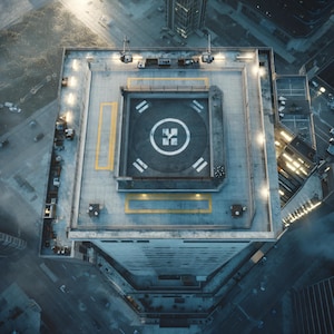 Könnte beinhalten: Luftaufnahme eines Wolkenkratzerdachs mit einem Hubschrauberlandeplatz. Der Landeplatz ist mit weißen und gelben Linien und einem zentralen Symbol markiert. Lichter und architektonische Details umgeben den Landeplatz. Aufnahme von oben.