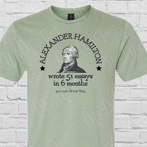 May include: A light green t-shirt with a black graphic of Alexander Hamilton's portrait in a circle with the text "Alexander Hamilton wrote 51 essays in 6 months you can finish this..."