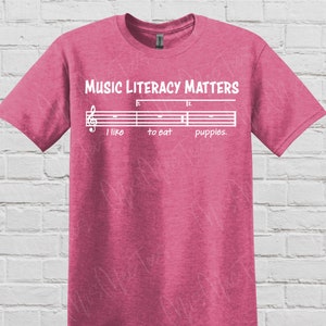 May include: Pink t-shirt with a white graphic that says "Music Literacy Matters" and features a musical staff with the words "I like to eat puppies."