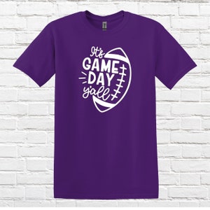 May include: Purple t-shirt with a white graphic of a football and the text "It's Game Day Y'all".