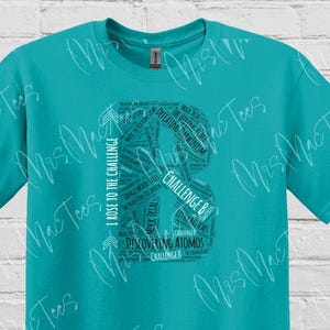 May include: A turquoise t-shirt featuring a large, stylized letter "B" filled with text. The text includes phrases like "I Rose to the Challenge" and "Discovering Atomos." The shirt has a crew neck and short sleeves.