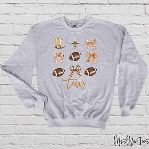 May include: A gray sweatshirt with a design featuring footballs, bows, boots, a longhorn steer, and the word "Texas".