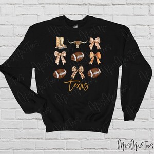 May include: Black sweatshirt with a design featuring a brown steer head, footballs, boots, and bows in brown and white plaid. The text "Texas" is written in orange.