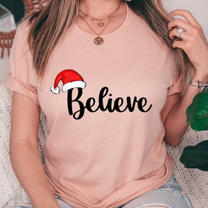 クリスマスを信じるシャツ、サンタクロースの帽子を信じるTシャツ、クリスマスの面白いギフトシャツ、クリスマスサンタシャツ、モチベーションを高める冬のホリデーシャツ
